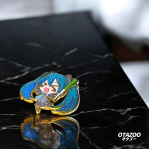 Miku pin brooch with blue glitter hair and gold plating, sitting on a black marble desk — a must-have for Miku enamel pin fans.