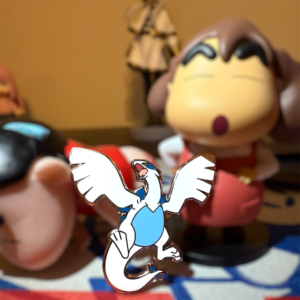 White and blue Lugia pin in a collector display scene with background anime toys